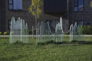 A light green railing is situated in the grass, located infront of a block of flats. The railing structure has an undulating top, with irregular spacing between the vertical bars.