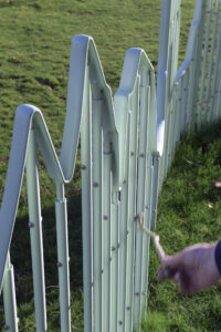 A close up image of a light green railing, with an undulating top can be seen with a hand drawing a stick along the vertical rails. 